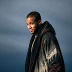 A man wearing a dark hooded jacket with a black sherpa collar and a mountain landscape print, photographed outdoors against a moody blue-gray sky.