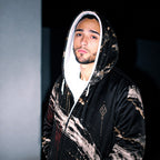 A man wearing a black hooded sweatshirt with a white and beige marble-like paint splatter pattern, featuring a white-lined hood and a small geometric logo on the chest.