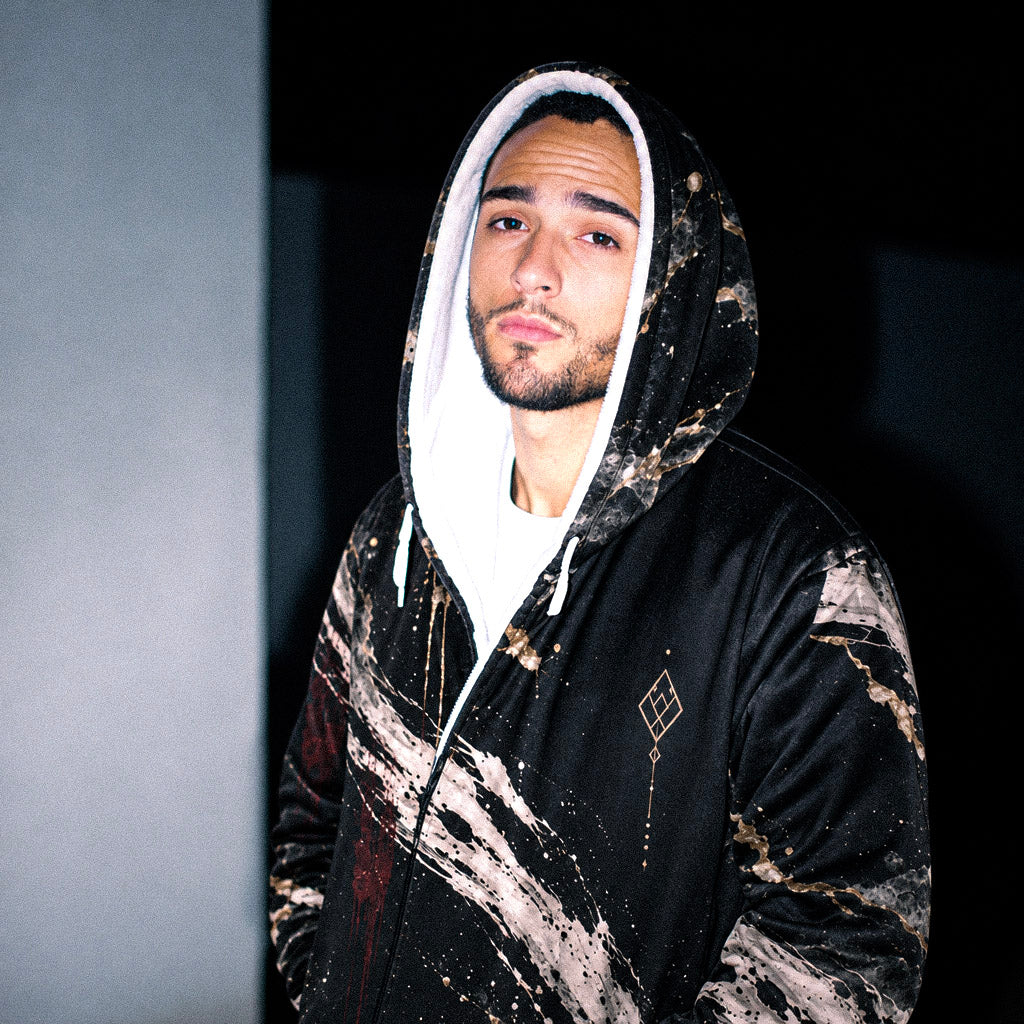 A man wearing a black hooded sweatshirt with a white and beige marble-like paint splatter pattern, featuring a white-lined hood and a small geometric logo on the chest.