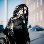 A person wearing a black hooded garment with a marbled, stone-like pattern in black, gray, and white, photographed outdoors on a blurred street background.