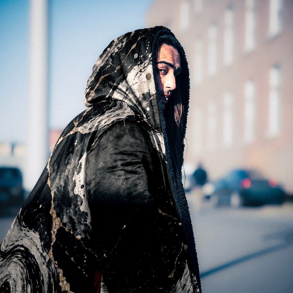 A person wearing a black hooded garment with a marbled, stone-like pattern in black, gray, and white, photographed outdoors on a blurred street background.