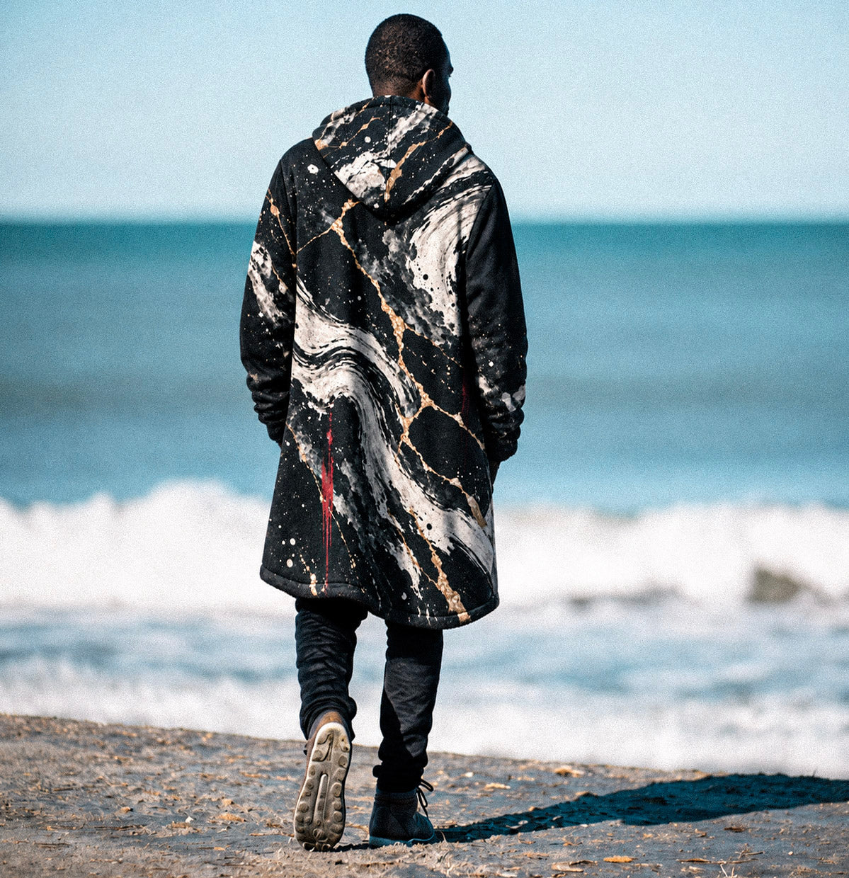 A man viewed from behind walking on a beach toward the ocean, wearing a long black hooded jacket with a white and gray marble pattern and subtle gold veining.