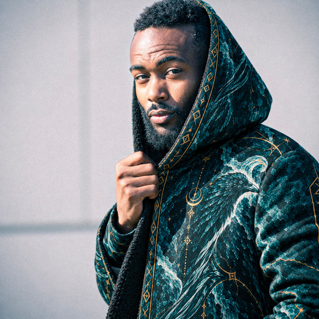 A man wearing a dark teal hooded robe with gold celestial and wave patterns, holding the hood up against a plain light gray wall.