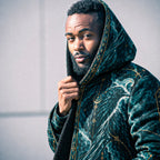 A man wearing a dark teal hooded robe with gold celestial and wave patterns, holding the hood up against a plain light gray wall.