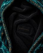 Close-up of a plush hooded blanket with a black sherpa-like interior and a teal patterned exterior featuring gold geometric and star motifs. A black brand patch reading RAVNVEIL is visible inside.