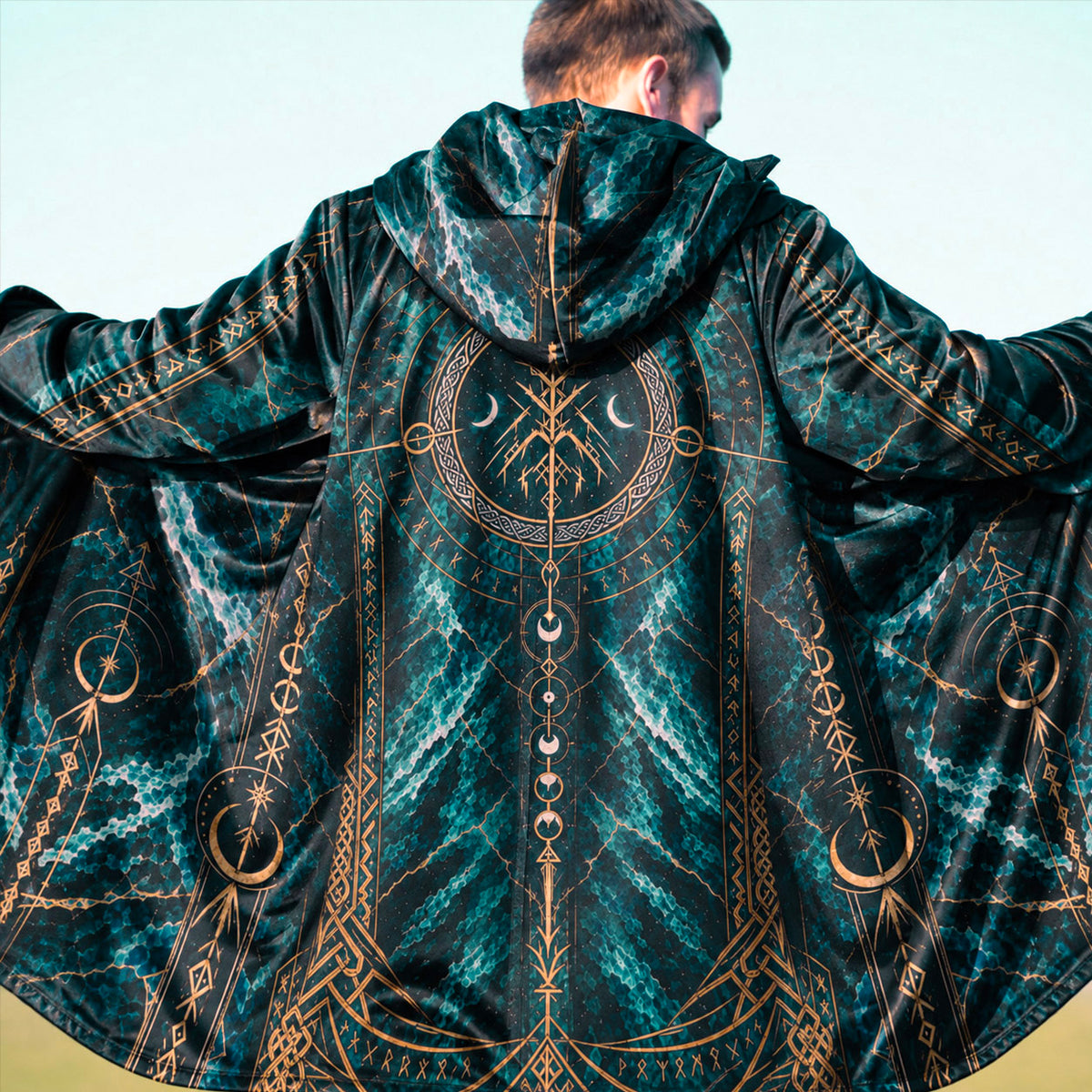 A man viewed from behind wearing a hooded robe with a dark teal and black marbled pattern and intricate gold celestial and geometric designs.
