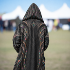 A person viewed from behind wearing a long black hooded robe or coat with ornate multicolored feather-like patterns, standing outdoors in a grassy field with blurred white tents in the background.
