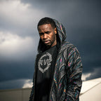 A man wearing a black t-shirt with a white graphic and a highly detailed, multicolored paisley-patterned hooded jacket, set against a dramatic dark stormy sky.