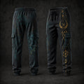 A pair of dark charcoal cargo jogger pants shown from the front and back against a dark studio background. The pants feature a subtle teal and gold cosmic nebula print, with gold celestial and geometric symbols printed on the left leg.