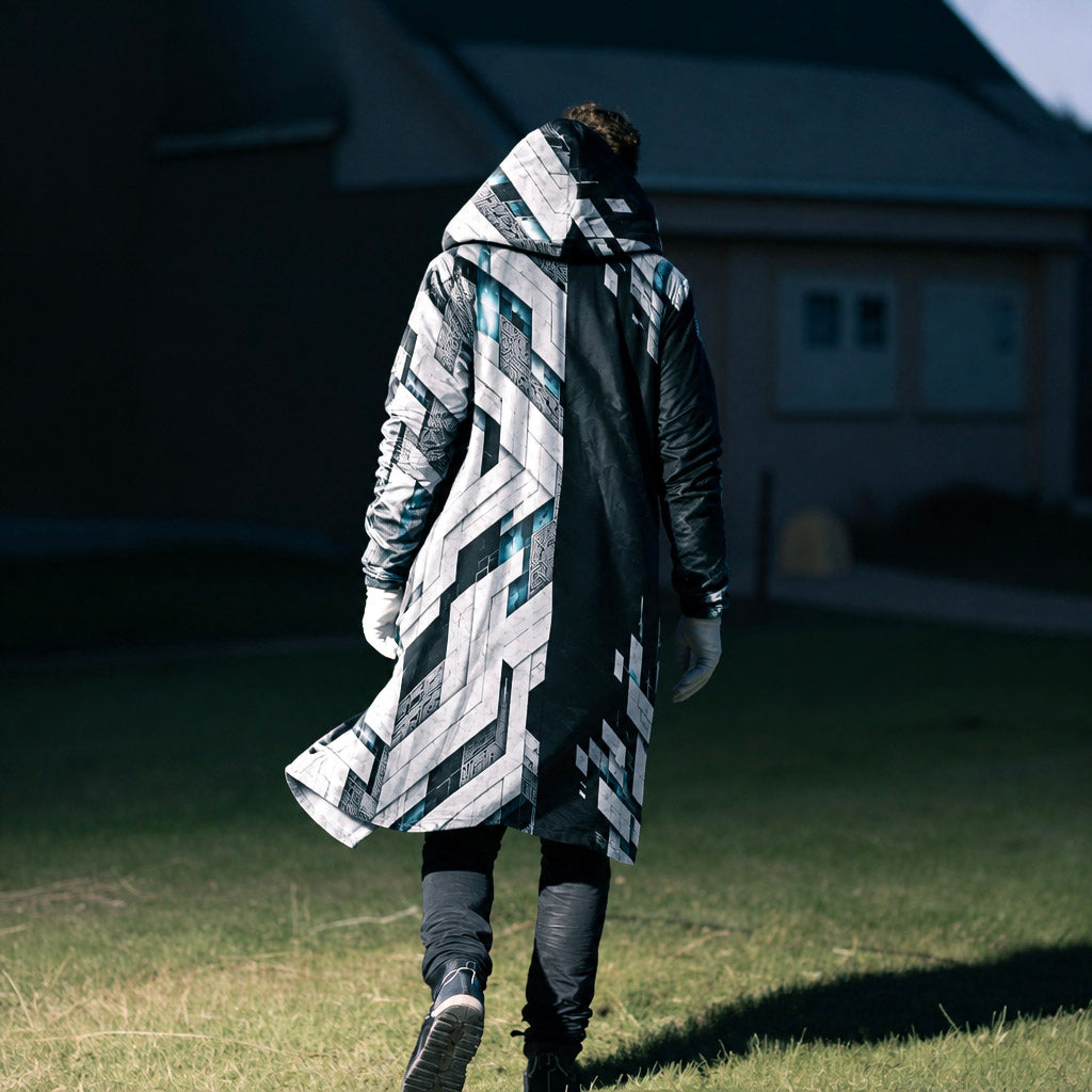 A person viewed from behind wearing a long hooded coat with a bold geometric black, white, and teal pattern, walking on a grassy lawn in front of a house.