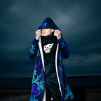 A person wearing a long hooded coat with a blue and purple fractal pattern over a black outfit, standing against a dark moody sky.