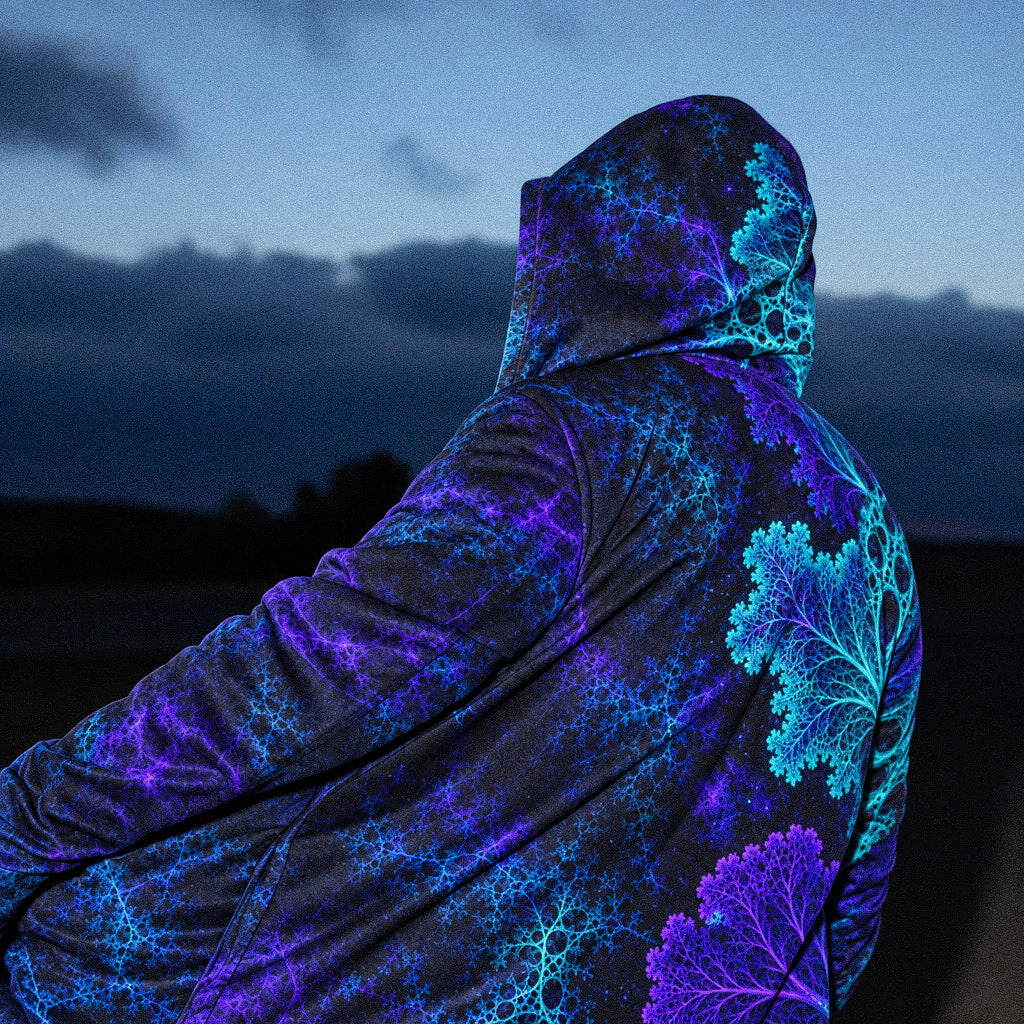 Voidbloom Eternal Cloak featuring a black base with vibrant blue, cyan and purple fractal patterns, shown from the back with the hood up in a dark outdoor setting.