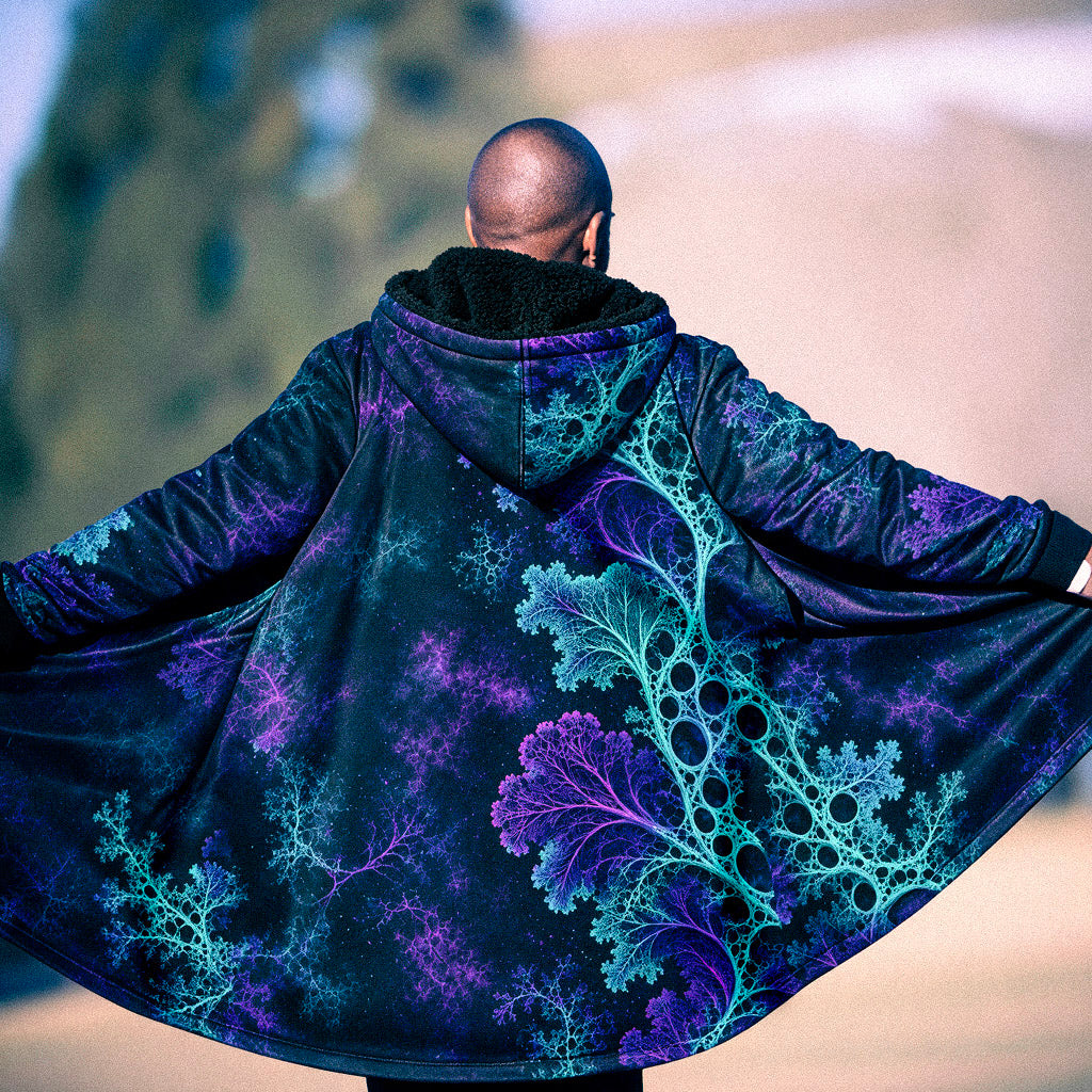 A man viewed from behind wearing a large open-front hooded coat with a dark base and vivid fractal-like branching patterns in purple and bright aqua. The background is a softly blurred outdoor scene.