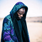 A man wearing a black hooded jacket with a vibrant teal and purple fractal pattern, standing in a desert landscape.