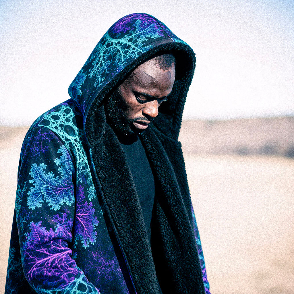 A man wearing a black hooded jacket with a vibrant teal and purple fractal pattern, standing in a desert landscape.