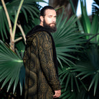 A bearded man wearing a black hooded robe with an intricate gold Norse-style knotwork and compass pattern, standing in front of a lush background of green palm leaves.